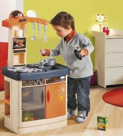 Un petit garçon est penché sur une casserole posée sur une cuisinière proportionnée à sa taille. Il tient le couvercle de la casserole dans la main gauche et une cuillère dans la droite, pour goûter sa préparation semble-t-il.